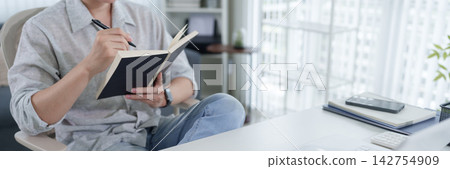 Asian woman writing notes in journal while working from home with bright window view and copy space Asian woman writing notes in journal while working from home with bright window view and copy space 142754909
