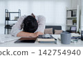 Exhausted young Asian professional resting head on desk surrounded by work materials in modern home office 142754940