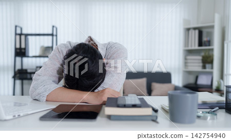 Exhausted young Asian professional resting head on desk surrounded by work materials in modern home office Exhausted young Asian professional resting head on desk surrounded by work materials in modern home office 142754940