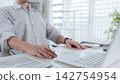 Asian professional typing on laptop at modern home office desk with documents and natural lighting 142754954