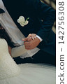 Groom in a suit with a boutonniere holds the hand of a bride in a white dress with pearls at a wedding. Romantic wedding moment, newlyweds holding hands. 142756308