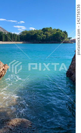 Turquoise coastal waters frame a distant pine covered islet in a calm sheltered bay Turquoise coastal waters frame a distant pine covered islet in a calm sheltered bay 142756536