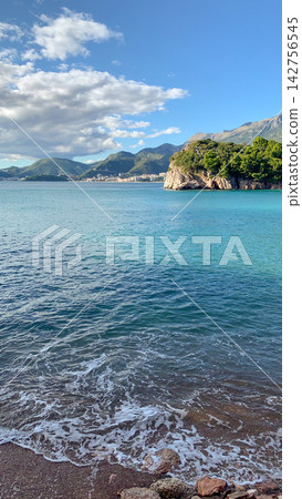 Turquoise waves rolling onto pebbles along rocky cliff base with pine trees swaying in coastal breeze under deep blue sky on sunny summer afternoon in Europe Turquoise waves rolling onto pebbles along rocky cliff base with pine trees swaying in coastal breeze under deep blue sky on sunny summer afternoon in Europe 142756545