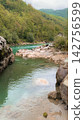 Mountain river featuring turquoise glacial water flows past jagged stone formations and boulders along a natural bank surrounded by dense forest slopes under a moody overcast sky lighting 142756599