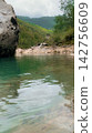 Large stone boulder sitting by a quiet turquoise mountain river with dense green forest background providing a grounded perspective of the serene riverside scenery during an overcast spring afternoon 142756609