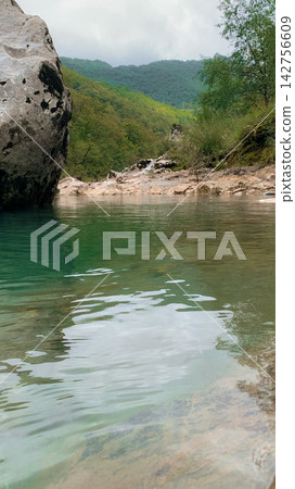 Large stone boulder sitting by a quiet turquoise mountain river with dense green forest background providing a grounded perspective of the serene riverside scenery during an overcast spring afternoon Large stone boulder sitting by a quiet turquoise mountain river with dense green forest background providing a grounded perspective of the serene riverside scenery during an overcast spring afternoon 142756609