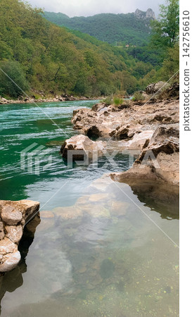 Jagged rocks lining the edge of a vibrant emerald river winding through dense forest hills during a peaceful spring day with soft natural light enhancing the clear water and canyon textures Jagged rocks lining the edge of a vibrant emerald river winding through dense forest hills during a peaceful spring day with soft natural light enhancing the clear water and canyon textures 142756610