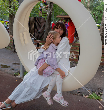 Mother embracing daughter inside circular playground seat with ropes course, trees, and wooden fencing behind them at outdoor adventure park, showing family affection, care, and leisure Mother embracing daughter inside circular playground seat with ropes course, trees, and wooden fencing behind them at outdoor adventure park, showing family affection, care, and leisure 142756761