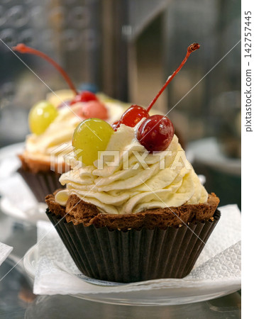Muffin shaped pastries in paper wrappers with cream on top, red cherries and a green grape are arranged in a confectionery display case, creating a colorful dessert presentation Muffin shaped pastries in paper wrappers with cream on top, red cherries and a green grape are arranged in a confectionery display case, creating a colorful dessert presentation 142757445