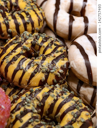 Fresh ring donuts with yellow glaze decorated with chocolate stripes and seeds arranged on a bakery display, featuring sweet baked pastries for customers Fresh ring donuts with yellow glaze decorated with chocolate stripes and seeds arranged on a bakery display, featuring sweet baked pastries for customers 142757493