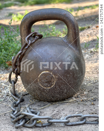 Heavy iron kettlebell with a chain attached standing on the ground at a shared outdoor gym area for bodybuilding, weight training and physical fitness activities Heavy iron kettlebell with a chain attached standing on the ground at a shared outdoor gym area for bodybuilding, weight training and physical fitness activities 142757544
