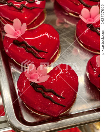 Red heart shaped pastries decorated with sweet pink flowers displayed in a pastry shop showcase, presenting an attractive and delicate selection of elegant desserts Red heart shaped pastries decorated with sweet pink flowers displayed in a pastry shop showcase, presenting an attractive and delicate selection of elegant desserts 142757696