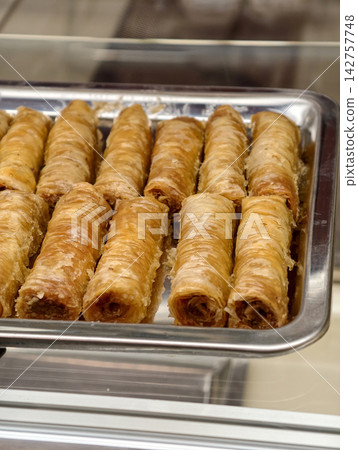 Sweet tube shaped baklava pastries presented in a pastry shop showcase, highlighting an attractive traditional dessert with delicate layers and a neat rolled appearance Sweet tube shaped baklava pastries presented in a pastry shop showcase, highlighting an attractive traditional dessert with delicate layers and a neat rolled appearance 142757748
