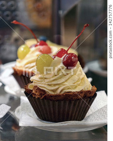 Muffin shaped pastries in paper wrappers feature a cream topping decorated with red cherries and a green grape, presenting an attractive selection of colorful sweet treats Muffin shaped pastries in paper wrappers feature a cream topping decorated with red cherries and a green grape, presenting an attractive selection of colorful sweet treats 142757776