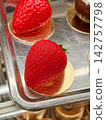 Bright red pastries made in the shape of strawberries displayed in a pastry shop showcase, offering an appetizing presentation of sweet treats with an elegant decorative appearance 142757798