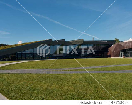 Architectural complex featuring angular glass facade, dramatic slanted rooflines, brick accents, reflective windows, and expansive landscaped grounds under clear blue sky 142758024