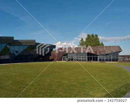 Modern glass facade building with expansive windows reflecting daylight, set against clear blue sky, with trees and green lawn Modern glass facade building with expansive windows reflecting daylight, set against clear blue sky, with trees and green lawn 142758027