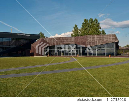 Modern architectural building with wooden lattice roof, glass walls, blue sky, and green lawn with trees Modern architectural building with wooden lattice roof, glass walls, blue sky, and green lawn with trees 142758028