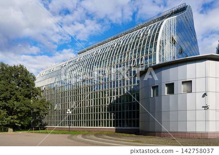 Modern glass facade building with slanted roof and large reflective windows under bright blue sky and white clouds, sleek downtown urban architecture in daylight Modern glass facade building with slanted roof and large reflective windows under bright blue sky and white clouds, sleek downtown urban architecture in daylight 142758037
