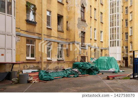 Yellow building facade with peeling plaster, weathered walls, exposed masonry, and construction clutter in sunlit urban courtyard depicting aged residential architecture and ongoing renovation Yellow building facade with peeling plaster, weathered walls, exposed masonry, and construction clutter in sunlit urban courtyard depicting aged residential architecture and ongoing renovation 142758070