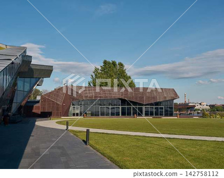 Contemporary glass and metal office building with angular facade, reflective windows, landscaped green lawn, paved walkway, tall trees, and bright blue sky Contemporary glass and metal office building with angular facade, reflective windows, landscaped green lawn, paved walkway, tall trees, and bright blue sky 142758112