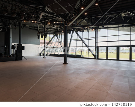Industrial event hall interior with exposed metal framework, ceiling rigging, hanging stage lights, and expansive glass wall revealing daytime city skyline panorama Industrial event hall interior with exposed metal framework, ceiling rigging, hanging stage lights, and expansive glass wall revealing daytime city skyline panorama 142758119
