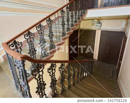 Intricate wrought iron staircase railing winds along warm peach plaster walls inside historic building, ornate balusters and curved wooden handrail frame stone steps, dramatic interior staircase scene 142758120