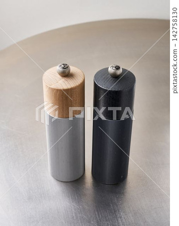 Pair of ribbed salt and pepper grinders, cylindrical metal bodies, standing upright on stainless steel surface with natural and dark wood heads, close-up view of knurled caps Pair of ribbed salt and pepper grinders, cylindrical metal bodies, standing upright on stainless steel surface with natural and dark wood heads, close-up view of knurled caps 142758130