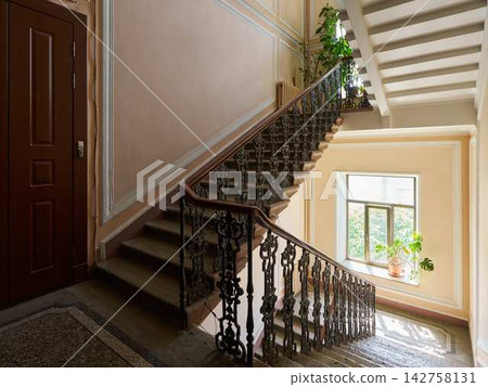 Grand indoor staircase with ornate wrought iron railing, peach paneled walls, large window with daylight, potted plant, warm sunlight, and vintage style 142758131