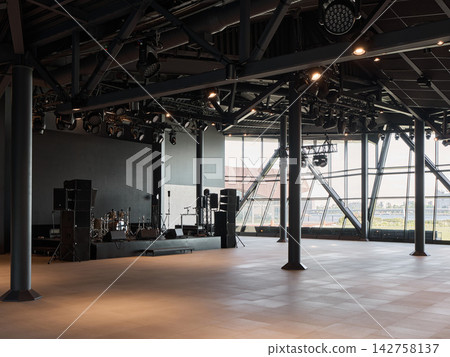 Interior concert hall stage featuring extensive lighting rig, steel beams and trusses, moody ambience, blank platform, and bright windows overlooking cityscape nighttime Interior concert hall stage featuring extensive lighting rig, steel beams and trusses, moody ambience, blank platform, and bright windows overlooking cityscape nighttime 142758137
