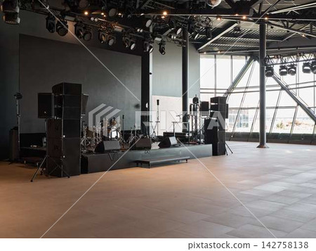 Concert stage inside modern arena during setup with expansive overhead lighting rig, large speakers, drum kit, microphones, and glass interior Concert stage inside modern arena during setup with expansive overhead lighting rig, large speakers, drum kit, microphones, and glass interior 142758138
