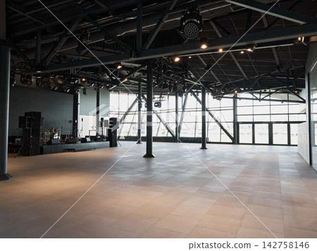 Industrial concert hall interior with elevated lighting rigs, heavy steel beams, exposed piping, and empty stage beneath expansive panoramic windows, showcasing modern architectural space 142758146