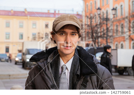 Man student looking at camera on the street during his break. Young guy in city wearing casual winter jacket and baseball cap. Urban lifestyle and education concept for youth. 142759061