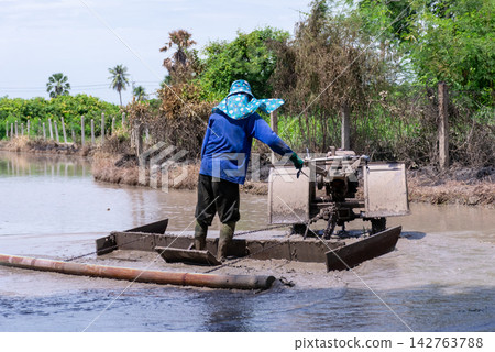 Farmer operates agricultural machinery muddy pond for soil preparation water management rural farming work surrounded by tropical vegetation Farmer operates agricultural machinery muddy pond for soil preparation water management rural farming work surrounded by tropical vegetation 142763788