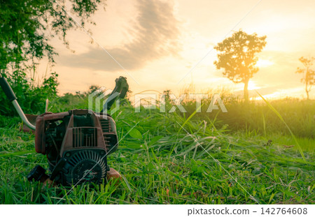 Portable petrol grass cutting machine resting in freshly cut green field at sunrise, agricultural equipment used by farmers for mowing vegetation, maintaining farmland and preparing livestock feed. Portable petrol grass cutting machine resting in freshly cut green field at sunrise, agricultural equipment used by farmers for mowing vegetation, maintaining farmland and preparing livestock feed. 142764608
