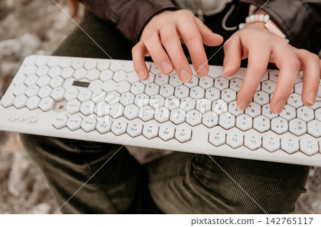 Typing keyboard hands. Person using a white hexagonal computer peripheral while sitting outdoors to work remotely Typing keyboard hands. Person using a white hexagonal computer peripheral while sitting outdoors to work remotely 142765117