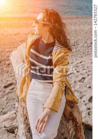 Woman beach jacket sits on a large driftwood log at the shore during a bright sunny afternoon with warm lens flare 142765327