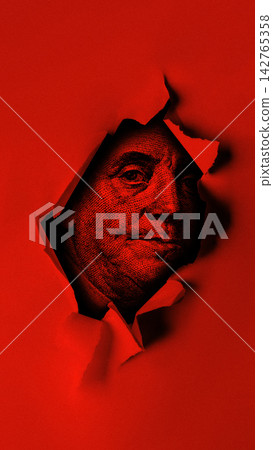 Close-up of a dollar bill peeking through a tear in red paper 142765358