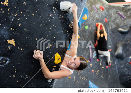 Sporty strong young female exercising in boulder climbing hall without rope, reaching new results, enjoying new challenges in amusement park Sporty strong young female exercising in boulder climbing hall without rope, reaching new results, enjoying new challenges in amusement park 142765411