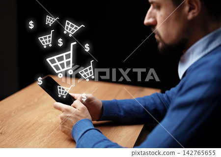 Man shops online using smartphone at a wooden table in a dark room with shopping cart icons Man shops online using smartphone at a wooden table in a dark room with shopping cart icons 142765655