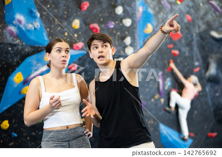 Positive young couple, male and female visitors discussing upcoming climbing artificial training rock wall in indoor bouldering gym Positive young couple, male and female visitors discussing upcoming climbing artificial training rock wall in indoor bouldering gym 142765692