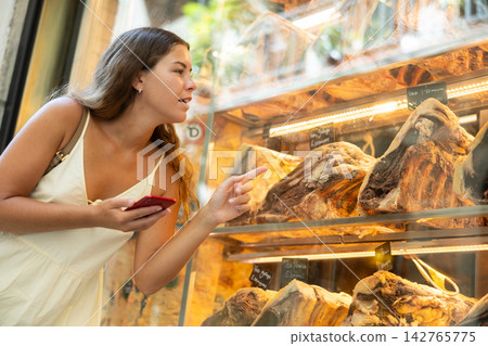 Girl with phone in hands looks at shop window, examines dried beef, looks at screen of smartphone Girl with phone in hands looks at shop window, examines dried beef, looks at screen of smartphone 142765775