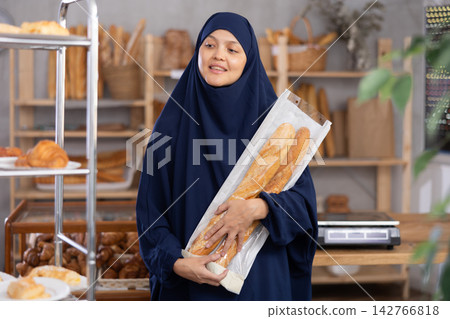 Middle-aged Muslim woman holding baguettes looks around in the bakery Middle-aged Muslim woman holding baguettes looks around in the bakery 142766818