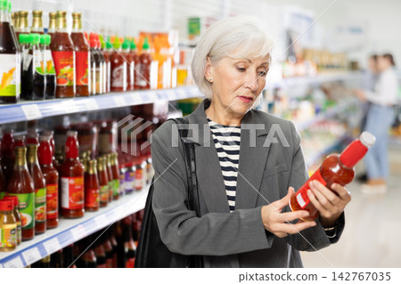 Senior woman customer is thinking about buying spicy and unusual red chilli tsuyu sauce. Senior woman customer is thinking about buying spicy and unusual red chilli tsuyu sauce. 142767035