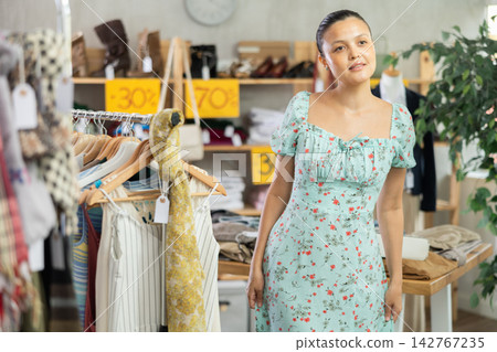 Asian woman chooses sundress in store and carefully examines her reflection in mirror Asian woman chooses sundress in store and carefully examines her reflection in mirror 142767235