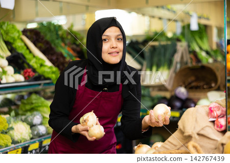 Portrait of positive female seller in hijab and apron puts onion on shelves in supermarket Portrait of positive female seller in hijab and apron puts onion on shelves in supermarket 142767439