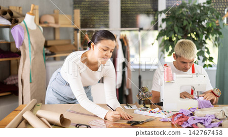 Attentive young man tailor sewing textile using sewing machine and female assistant drawing pattern on sheet of paper using curves in fashionable sewing atelier Attentive young man tailor sewing textile using sewing machine and female assistant drawing pattern on sheet of paper using curves in fashionable sewing atelier 142767458
