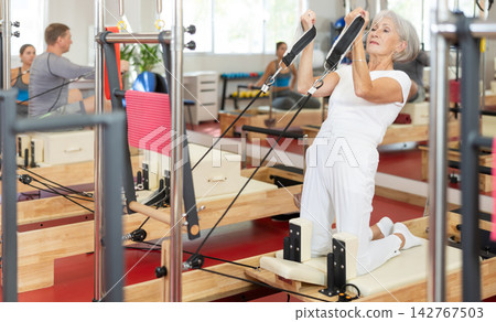 Sports club senior woman client performs exercise on reformer with cable and rope for arms Sports club senior woman client performs exercise on reformer with cable and rope for arms 142767503