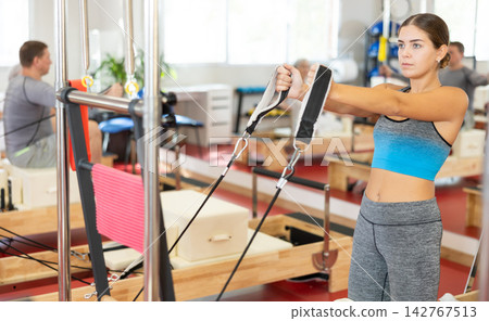 Sports club girl client on reformer with cable and rope performs exercise to stretch muscles of arms Sports club girl client on reformer with cable and rope performs exercise to stretch muscles of arms 142767513