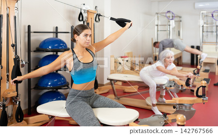 Sports club girl client on reformer with cable and rope performs exercise to stretch muscles of arms Sports club girl client on reformer with cable and rope performs exercise to stretch muscles of arms 142767566